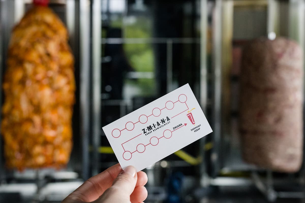a hand holding a white Kebap business card Change, with two meats on the grill in the background